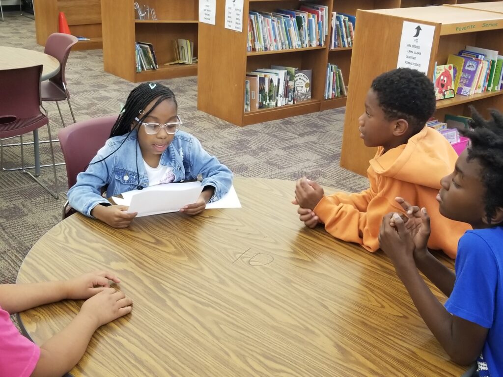 A young girl reads from a paper in a library setting while other campers listen attentively at the Greenhouse Communication Initiative summer camp.