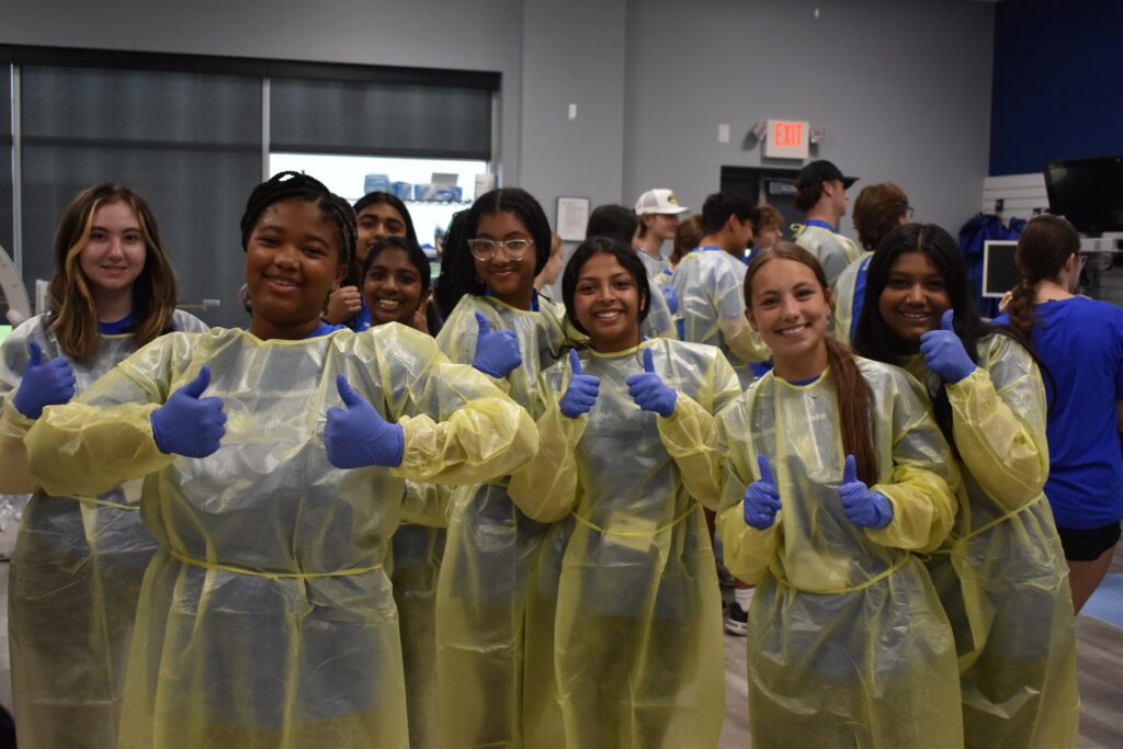 A group of teen girls wearing scrubs and medical gloves gives a thumbs up while participating in the Summer at SLU summer camp.