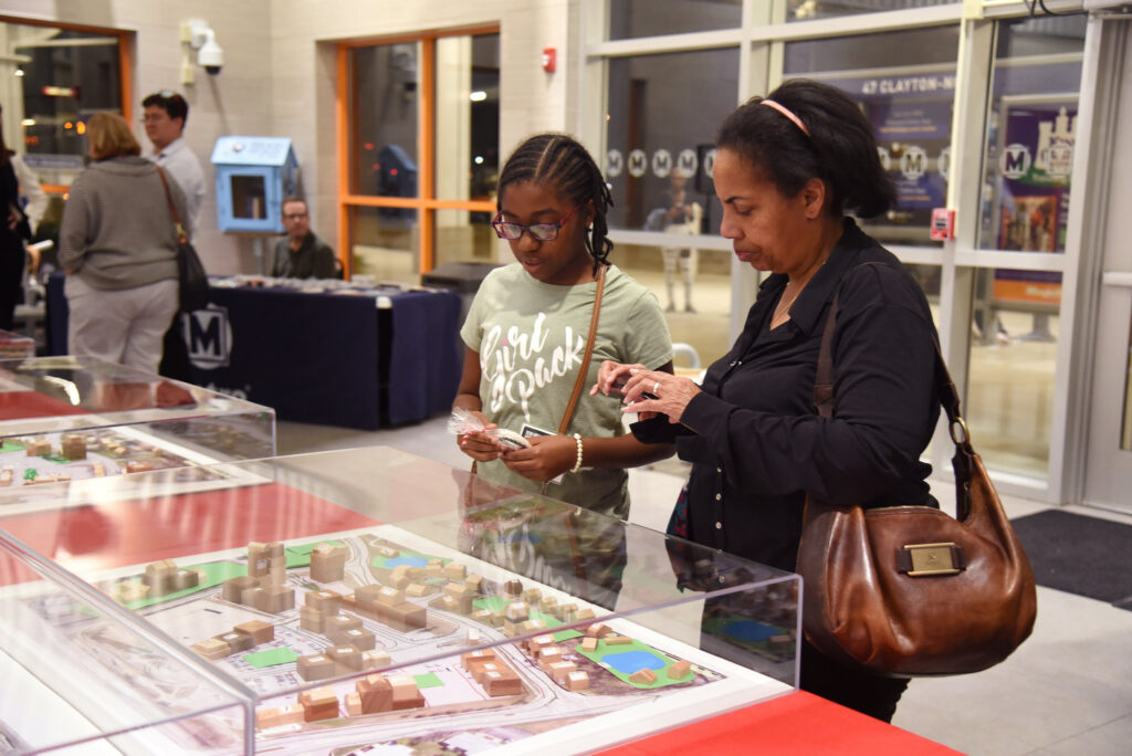 A tween girl and her mom look down at a glass case on a table. Inside the case is an architecture model with many small buildings created out of cardboard and paper. 