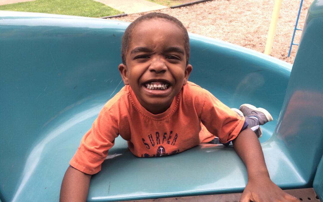 A child of about 3 or 4 years old is laying at the top of a blue plastic playground slide. He looks like he is about to slide down backwards. He has a huge grin on his face.