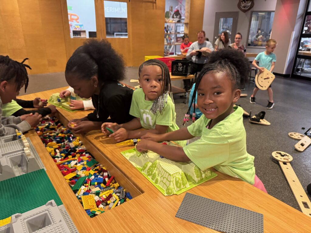 Two girls in matching green t shirts smile as they play with lego blocks at the Wesley House camp. Other kids in the room play and interact with wooden building pieces on the floor.