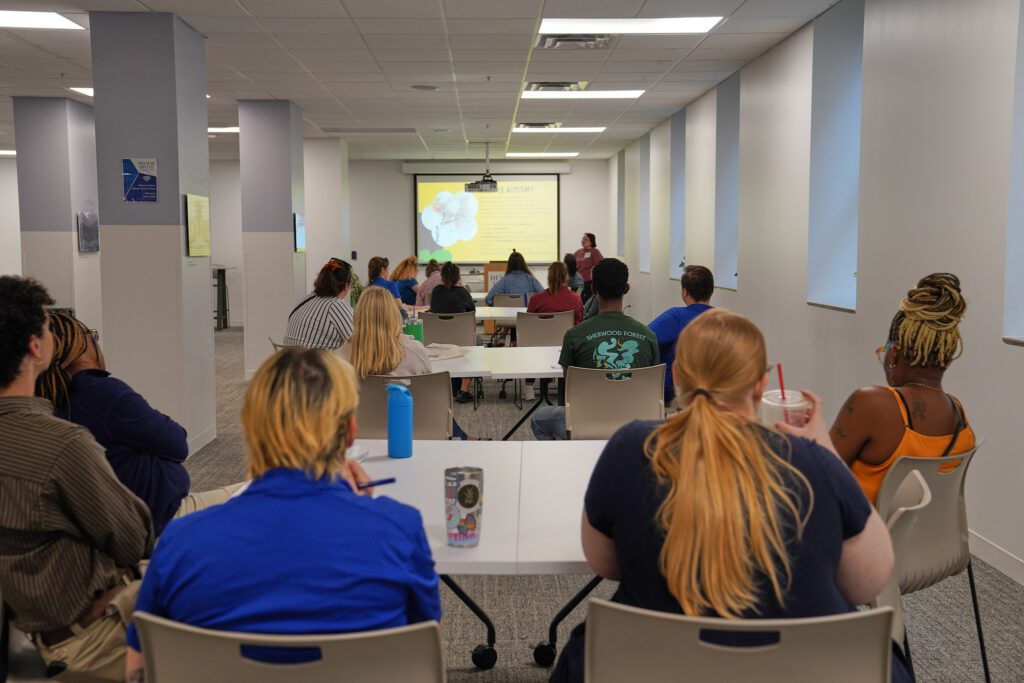 Camp providers sit at rows of tables to listen to a speaker on supporting neurodivergent campers. They are looking at a powerpoint slide titled what is autism? 