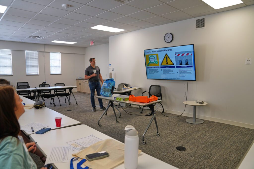 Providers watch a speaker from the National Weather service give a presentation on preparing for weather events during summer camp. The powerpoint slide reads Forecast and Hazards.
