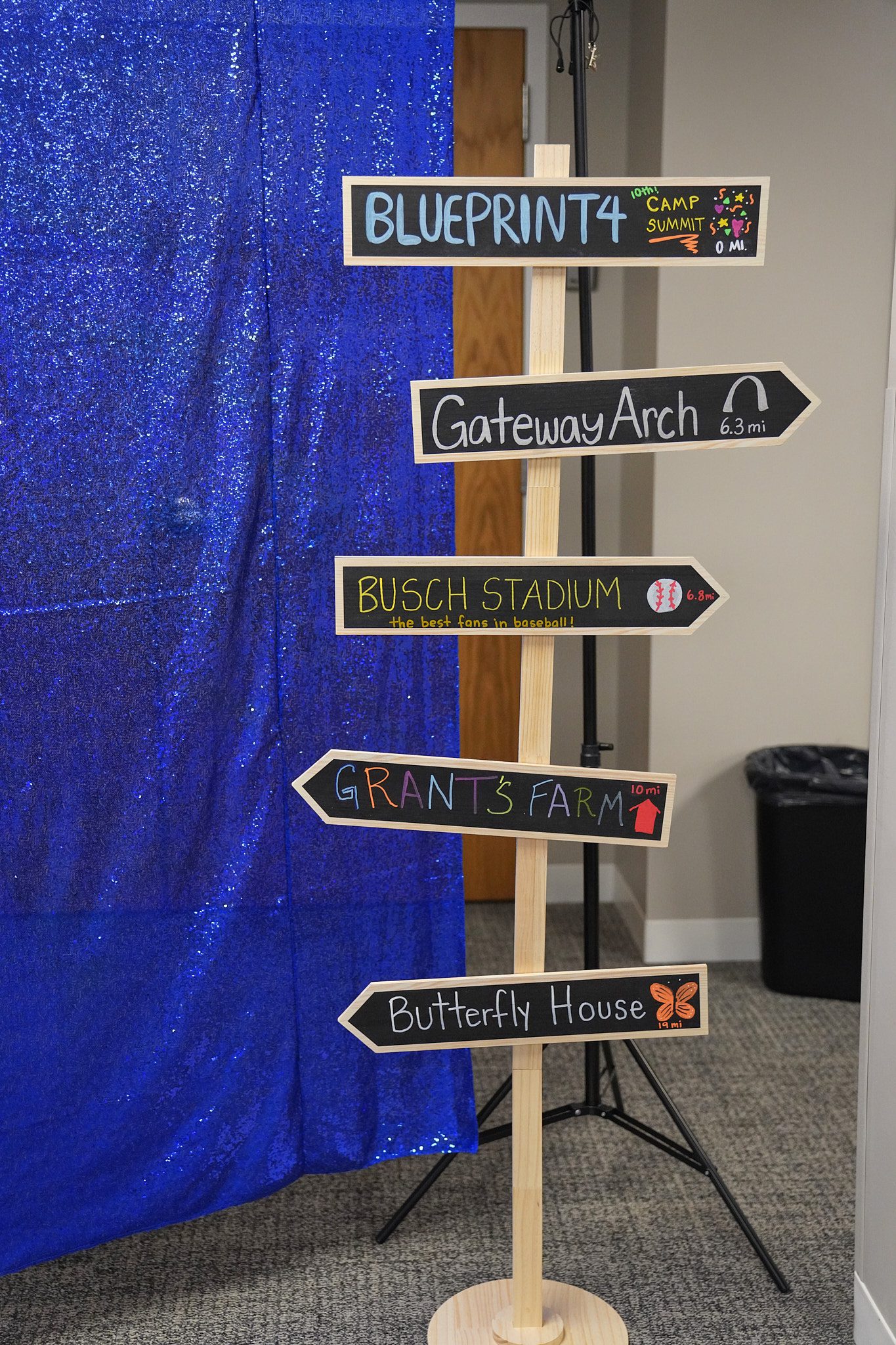 A directional sign with handwritten directions stands in front of a sparkly blue photo backdrop. The signs point to different well-known St. Louis landmarks.