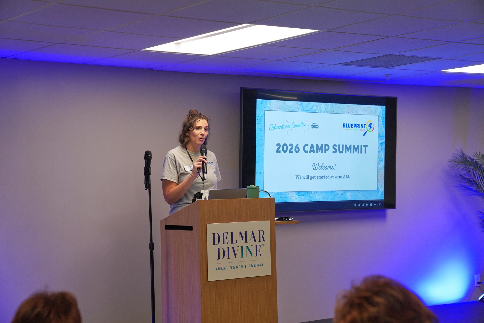 Program Manager Abbey Barrow gives a presentation at a podium branded with the Delmar Divine logo. A screen behind her reads 2026 Camp Summit.