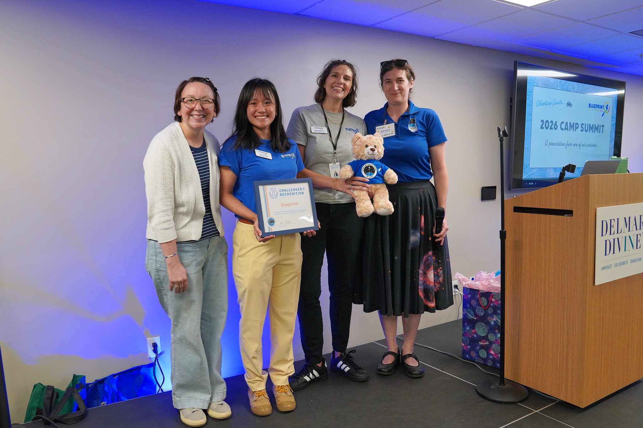 Blueprint4 staff pose for a photo with a camp provider from Challenger Learning Center. They are holding a stuff bear and a plaque.